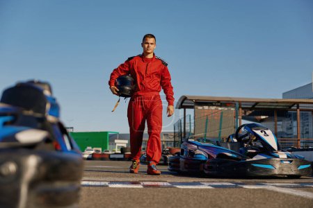 Photo for Equipped go-kart driver wearing overalls and holding helmet walking to car before race at starting line on motor racing track. Automotive sports activity, carting championship - Royalty Free Image