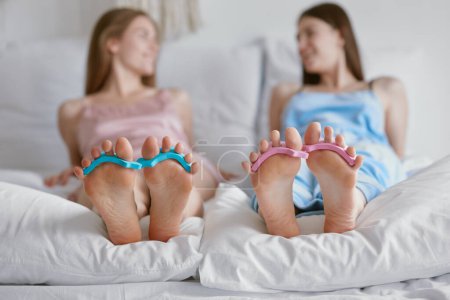 Photo for Closeup feet with toe spacers for nails painting. Two young woman friends lying in bed and gossiping having nice conversation. Sleepover party, family bonding - Royalty Free Image