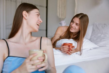 Téléchargez les photos : Deux jeunes femmes souriantes bavardant tout en buvant du thé ou du café profitant d'une soirée pyjama. Concept d'amitié et de temps ludique ensemble - en image libre de droit