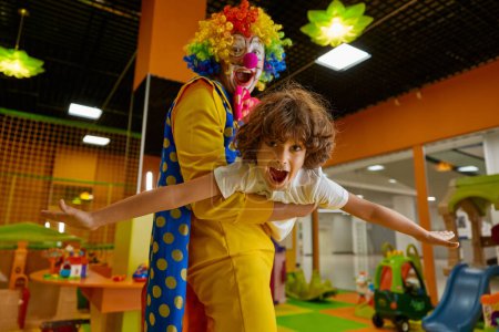 Photo for Joyful clown and kid feeling excitement playing game together at daycare center. Cheerful animator in colorful wig and costume holding happy screaming boy in hands - Royalty Free Image