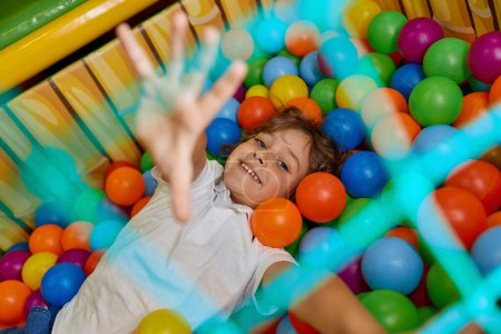 Téléchargez les photos : Enfant garçon heureux jouant dans le bassin rempli de boule en plastique multicolore. Enfant joyeux excité s'amusant dans une grande pataugeoire sèche à la salle de jeux intérieure - en image libre de droit