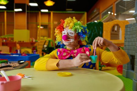 Téléchargez les photos : Drôle de clown portant une perruque multicolore et un costume jouant avec un ensemble de thé jouet à la salle de jeux pour enfants. Anniversaire fête animation spectacle concept - en image libre de droit