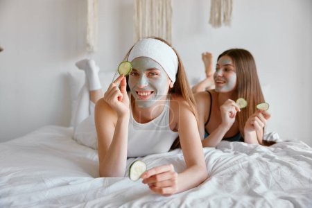 Téléchargez les photos : Portrait de deux amies célébrant la fête du spa à la maison en appliquant des tranches de concombre comme des patchs et un masque facial en argile cosmétique. Concept beauté et soins de la peau - en image libre de droit