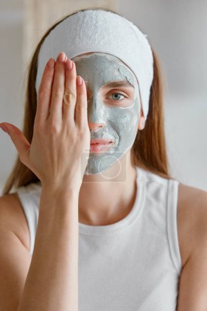 Foto de Retrato de mujer joven aplicando máscara de cosmetología facial de arcilla cubriendo media cara con la mano. Piel y belleza saludables, procedimiento de spa en casa con concepto de cosmética natural - Imagen libre de derechos