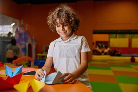 Foto de Retrato de niño feliz haciendo juguetes de papel de origami en la sala de juegos cubierta. Clase creativa y entretenimiento de hobby para niños talentosos en el parque infantil o guardería de la ciudad - Imagen libre de derechos