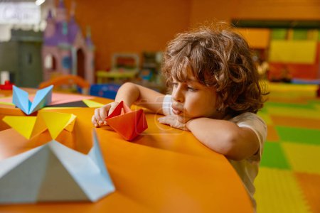 Foto de Niño lindo jugando barco de papel hecho a mano en la sala de juegos del centro de cuidado infantil. Entusiasta niño varón construyendo juguetes de origami disfrutando de la actividad de ocio en el área de diversión - Imagen libre de derechos