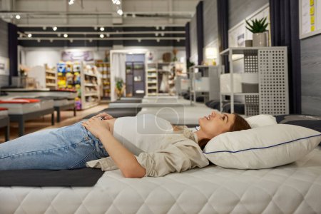 Photo for Young woman shopper choosing new bed at modern furniture showroom. Attractive female customer testing comfortable bedstead enjoying shopping activity on weekend - Royalty Free Image