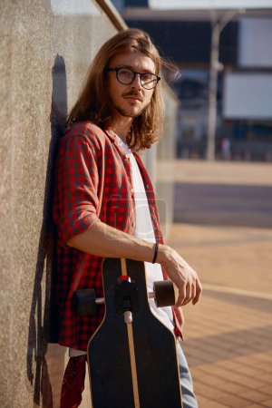 Téléchargez les photos : Portrait de taille de jeune homme hipster confiant debout avec longboard penché dos au mur et regardant la caméra avec un visage sérieux - en image libre de droit