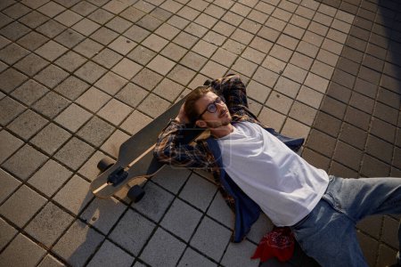 Téléchargez les photos : Hipster gars couché sur skateboard après l'entraînement de détente au coucher du soleil vue grand angle. Planche à roulettes passe-temps et temps libre des jeunes - en image libre de droit