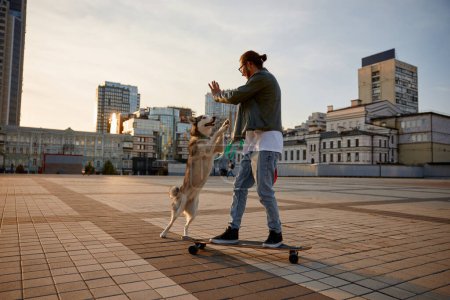 Téléchargez les photos : Élégant hipster guy en vêtements décontractés skateboard et chien de promenade se déplaçant ensemble à travers la place de la ville. Propriétaire d'animal de compagnie et chiot aimant profiter de l'activité physique lors d'une soirée calme au centre-ville - en image libre de droit