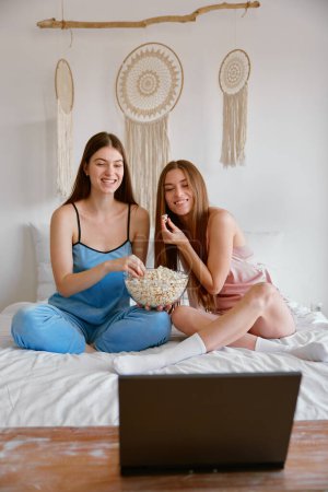 Photo for Excited young girls friends or sisters watching funny movie on laptop computer eating popcorn in bedroom. Pajama oversleep party concept - Royalty Free Image