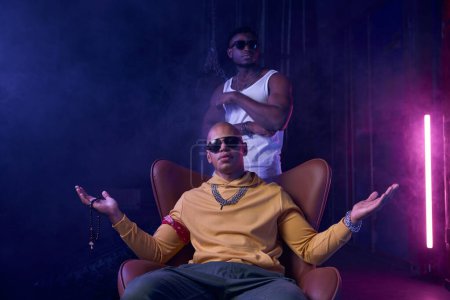 Photo for Two cool gangsters rap singers entertainment over industrial background. Young masculine crime boss sitting on leather armchair with bodyguard on backdrop - Royalty Free Image