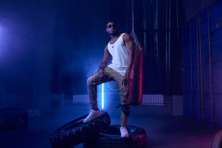 Moda de moda muscular rapero con gafas de sol de pie en luces de neón de club nocturno fresco con escenario decorado con saco de boxeo y neumático de rueda