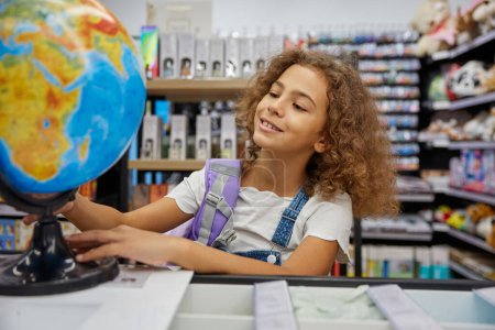 Photo for Little girl playing with school earth globe while shopping education stationery. Positive female student preparing for studying. Purchases with sale discount - Royalty Free Image