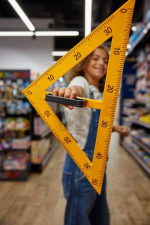 School girl student holding huge triangle ruler stationery in hands while standing at store. Creative studying tools for education
