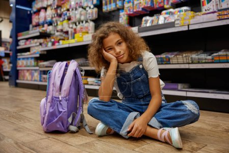 Bored or tired girl child sitting on floor of stationery shop. Cute little female student feeling exhausted after shopping to prepare for studying