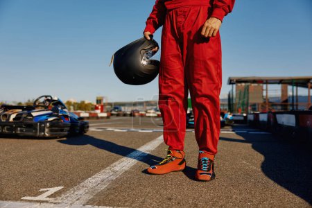 Cropped closeup shot of go-kart driver wearing overalls holding helmet in hands standing over motorsport training track