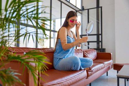 Foto de Mujer joven aplicando parche para los ojos mirando el espejo mientras está sentado en el sofá en la acogedora sala de estar. Inicio procedimiento de belleza - Imagen libre de derechos