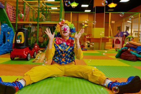 Photo for Funny childish clown feeling excitement and happiness playing with flying soap bubbles while sitting at playroom - Royalty Free Image