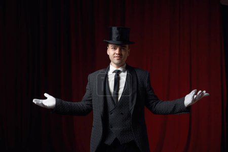 Photo for Young friendly smiling fashionable showman or illusionist in tailcoat, white gloves and top hat on stage greeting audience spreading arms while standing on theatre stage. Magic talent show start - Royalty Free Image