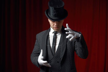 Téléchargez les photos : Portrait taille haute d'un bel homme magicien montrant un mystérieux tour de passe-passe agitant les mains dans des gants blancs sur scène. Spectacle magique et enchantement du public - en image libre de droit
