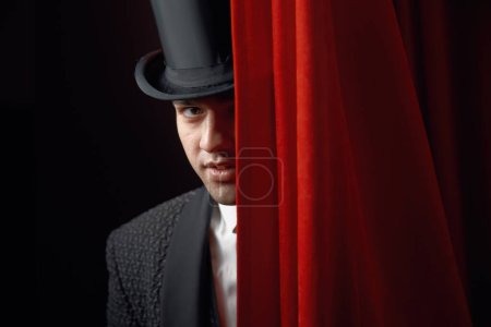 Der gut aussehende Magier lugt hinter dem Bühnenvorhang hervor. Junger männlicher Illusionist in elegantem Frack und Zylinder führt Zaubershow auf und überrascht das Publikum