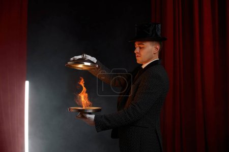 Téléchargez les photos : Magicien masculin portant costume de scène montrant tour avec illusion de feu magique avec plateau en acier. Performance mystérieuse avec un équipement magique - en image libre de droit