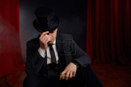 Photo for Elegant man magician wearing top hat and tuxedo suit holding magic cards in hands sitting on theatre stage with red drapery and mysterious smoke. Fantasy gambling show entertainment concept - Royalty Free Image