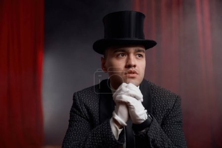 Téléchargez les photos : Magicien homme élégant portant chapeau haut de forme et costume de smoking sur scène avec draperie rouge et fumée mystérieuse. Fantasy show concept de divertissement - en image libre de droit