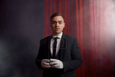 Photo for Portrait of handsome young male magician wearing suit standing with cane on stage decorated red velvet curtain - Royalty Free Image
