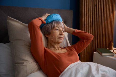 Femme âgée avec des maux de tête ou migraine tenant le sac avec de la glace se sentant malade, bouleversé et malheureux couché dans son lit. Concept de maladie chez les personnes âgées