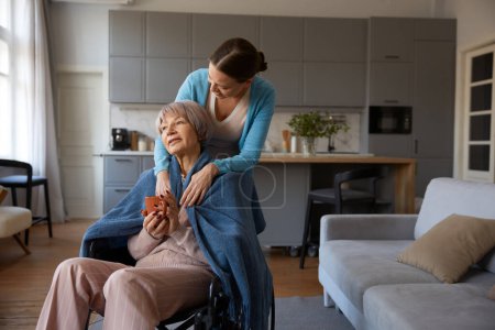 Photo for Loving adult daughter hugging old mother with disability sitting in wheelchair over cozy home interior. Happy family supporting relationship and bonding concept - Royalty Free Image