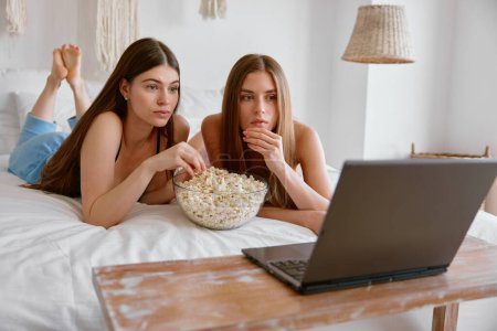 Téléchargez les photos : Jeunes filles amis ou sœurs regarder film drôle sur ordinateur portable manger du pop-corn dans la chambre. Pyjama concept de soirée pyjama - en image libre de droit