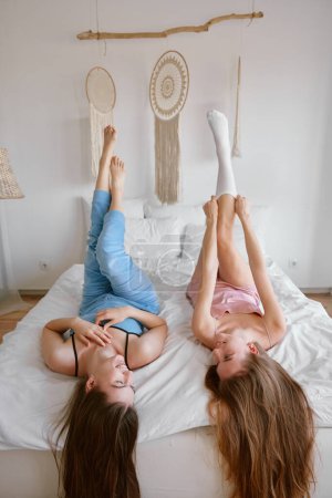 Photo for Two young beautiful girl friends wearing pajamas enjoying free time together lying on bed in bedchamber with raised up legs. Friendship and gossiping during slumber party celebration concept - Royalty Free Image
