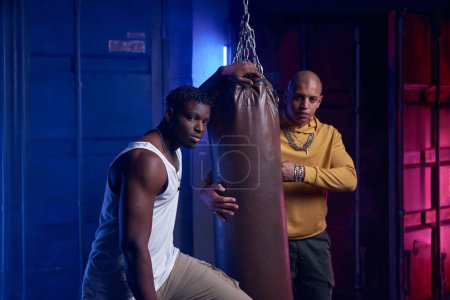 Photo for Young gang boys training with punching bag in garage. Two hipster guys friends exercising pumping muscles together - Royalty Free Image