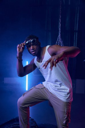 Der muskulöse Rapper mit Sonnenbrille steht im Neonlicht eines coolen Nachtclubs mit Bühne, die mit Boxsack und Radreifen dekoriert ist