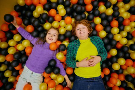 Foto de Encantadores niños divertidos tumbados en la piscina seca con bolas de plástico de colores disfrutando de un tiempo de diversión activa en el patio interior. Feliz infancia y vacaciones emocionadas - Imagen libre de derechos