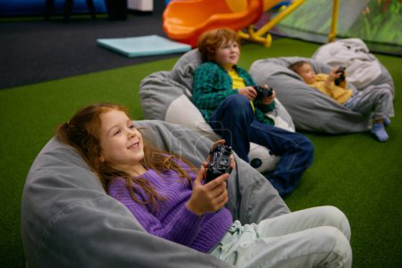 Téléchargez les photos : Petits enfants jouant à un jeu vidéo reposant sur des chaises douces après des jeux actifs dans le centre de divertissement - en image libre de droit