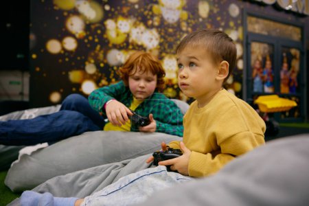 Foto de Niños jugando videojuegos descansando en sillas blandas después de juegos activos en el centro de entretenimiento - Imagen libre de derechos