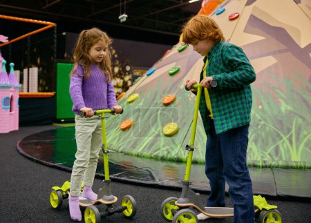 Photo for Carefree preschool children riding scooters together on indoor playground. Safety attraction equipment in game center for kids relaxation - Royalty Free Image