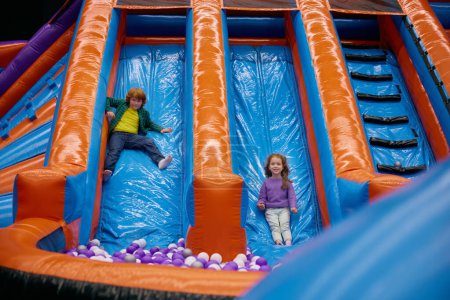 Téléchargez les photos : Portrait des enfants heureux excités sautant vers le bas sur la glissière gonflable. Loisirs amusants en sécurité aire de jeux pour enfants et enfance active - en image libre de droit