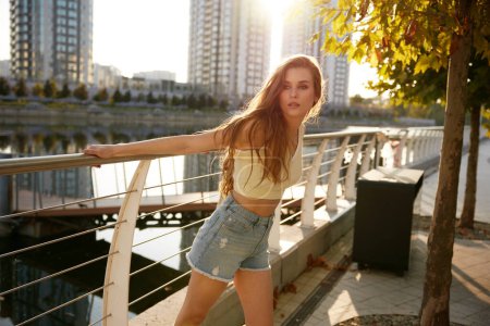 Téléchargez les photos : Charmante jeune femme en tenue tendance décontractée posant au coucher du soleil sur le parc de la ville près de la rive de la rivière. Heureux adolescent temps de loisirs - en image libre de droit