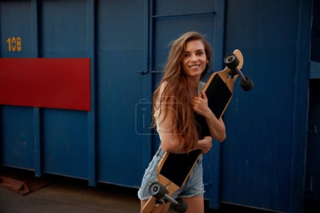 Téléchargez les photos : Joyeux sourire adolescent femme appréciant le skateboard son passe-temps sportif préféré. Jeune adolescente avec tenue décontractée tenant longboard dans les câlins - en image libre de droit