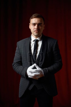 Téléchargez les photos : Portrait de beau jeune magicien masculin portant costume debout avec canne sur scène décoré rideau de velours rouge - en image libre de droit