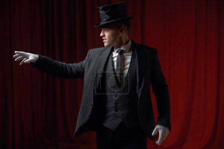 Photo for Young friendly fashionable showman or illusionist in tailcoat, white gloves and top hat on stage greeting audience spreading arms while standing on theatre stage. Magic talent show start - Royalty Free Image