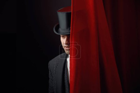 Apareciendo hombre guapo mago asomándose por detrás de la cortina de cortinas del escenario. Joven ilusionista masculino en elegante frac y sombrero de copa realizando espectáculo de magia y haciendo sorpresa para el público