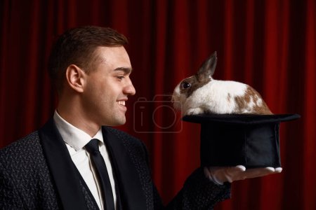 Foto de Retrato de vista lateral de hombre mago mirando conejo apareció en sombrero. Ilusionista profesional enfocándose con animales de compañía actuando en el escenario del teatro. Encantamiento y concepto de imaginación - Imagen libre de derechos