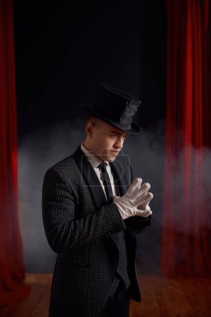 Photo for Elegant man magician wearing elegant suit standing in smoke on stage looking at audience. Young showman entertainer against red drapery curtain side view - Royalty Free Image