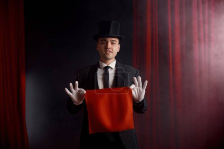 Photo for Young male magician actor showing trick with napkin in mysterious atmosphere of theatre stage with smoke. Illusion show and people professional occupation - Royalty Free Image