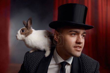 Retrato de hombre mago con conejo en el hombro sobre el escenario decorado con cortinas. Ilusionista profesional en traje de escena y sombrero de copa entretenido con conejito esponjoso animal de compañía